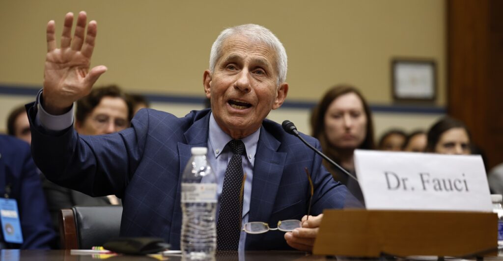 Anthony Fauci raises his hand while sitting down at a table with a water bottle next to him.