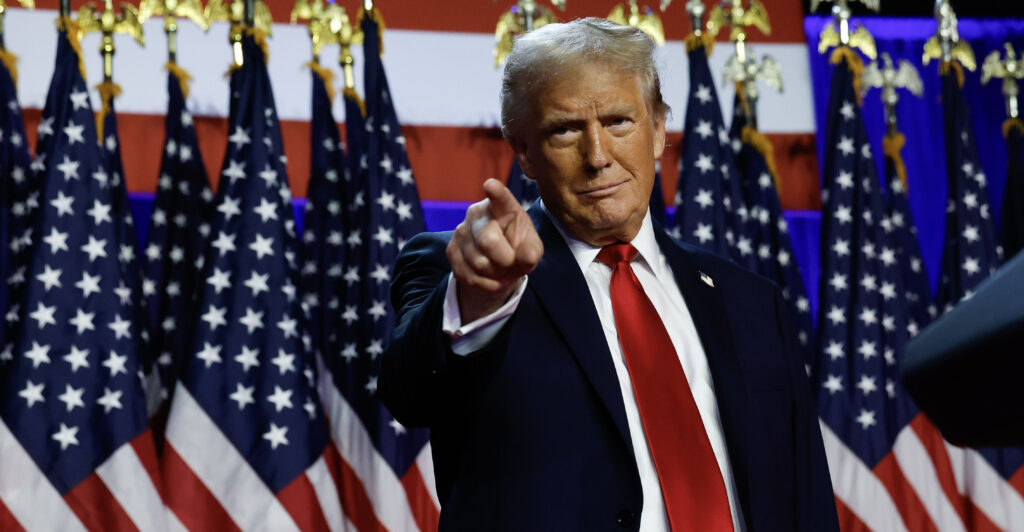 Donald Trump, wearing a black suit and red necktie, points to the crowd at a campaign event.
