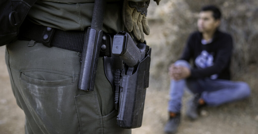 A U.S. Border Patrol agent, dressed in a green uniform with a black holstered gun, stands near a detainee, sitting in a black shirt.