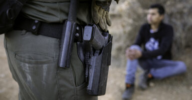 A U.S. Border Patrol agent, dressed in a green uniform with a black holstered gun, stands near a detainee, sitting in a black shirt.