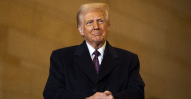 Donald Trump stands wearing a black coat with his hands clasped.