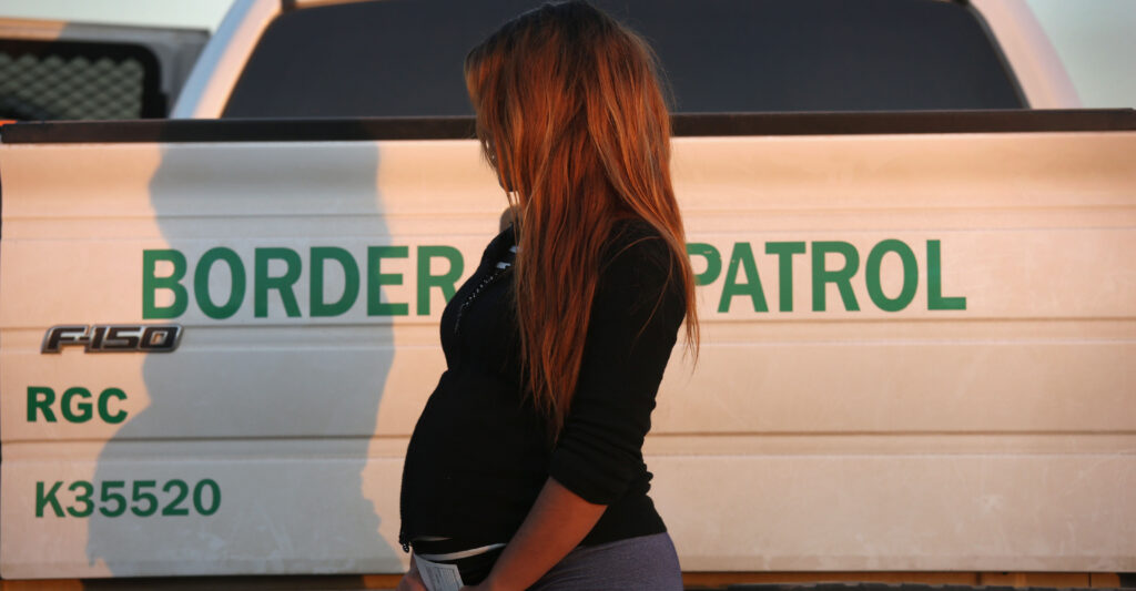 A pregnant women stands in a black shirt next to a white U.S. border patrol truck.