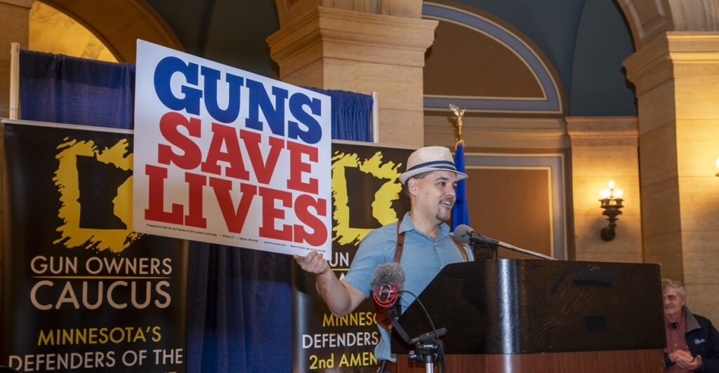 A man wearing a hat holds a sign that states, "Guns Save Lives."