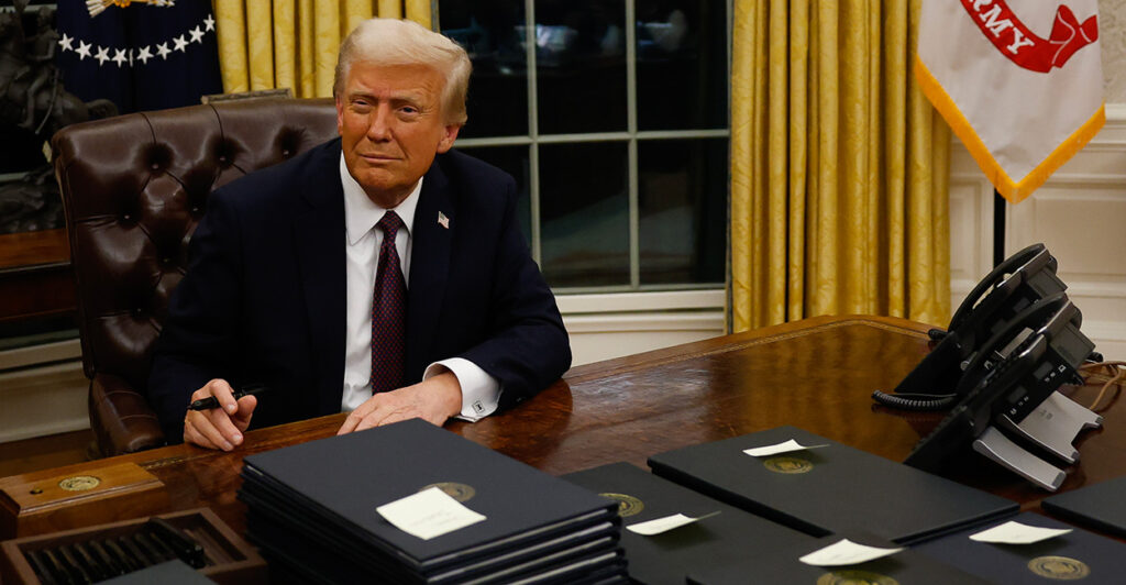 Donald Trump sits at his desk in a dark suit with stacks of executive orders.