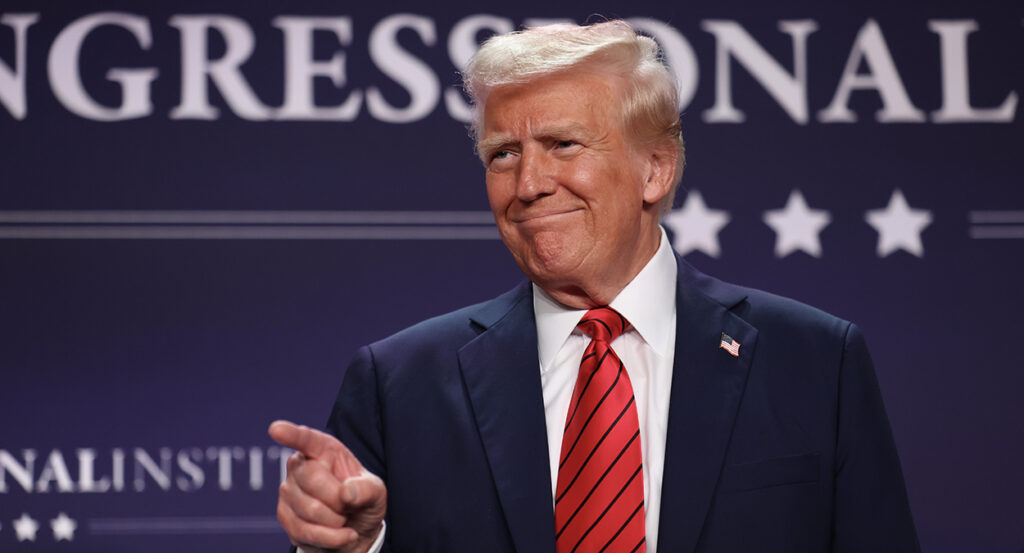 President Donald Trump points and smiles while wearing a blue suit with a red tie
