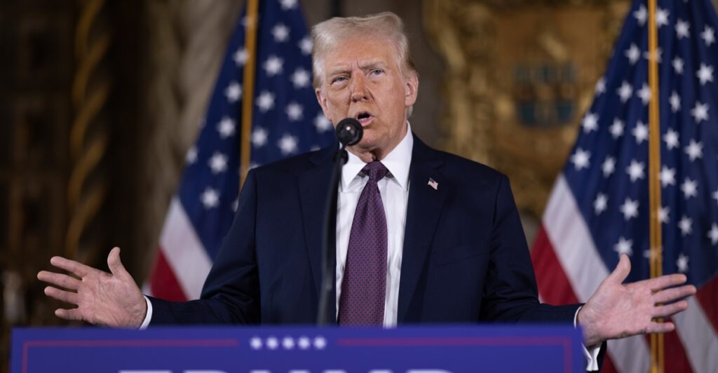 Donald Trump stands at a podium with his arms held out with American flags in the background.