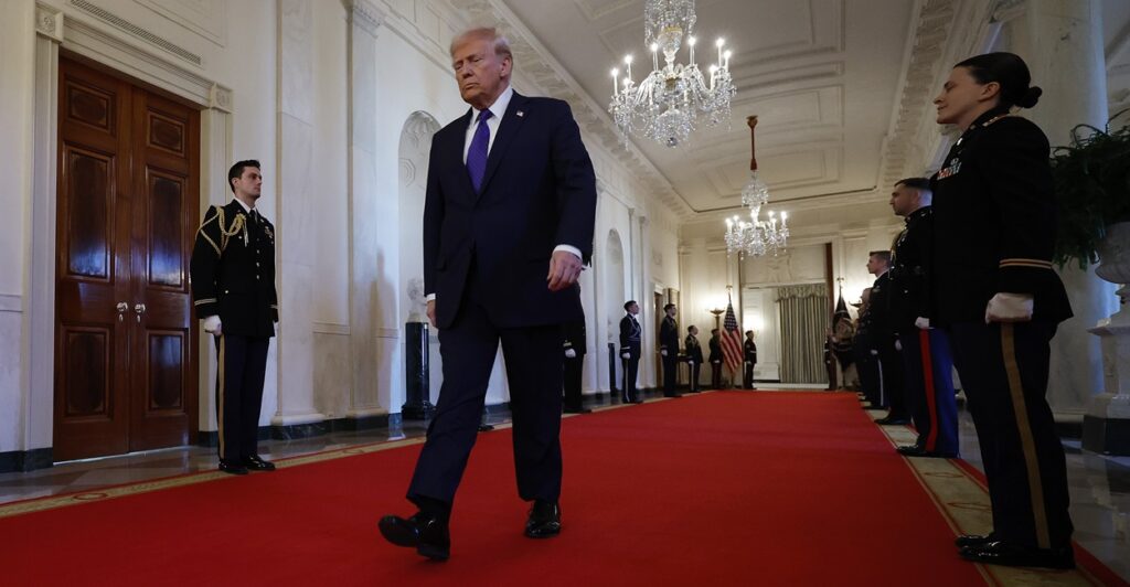 Donald Trump walks with his hands to his side in the White House on a red carpet.