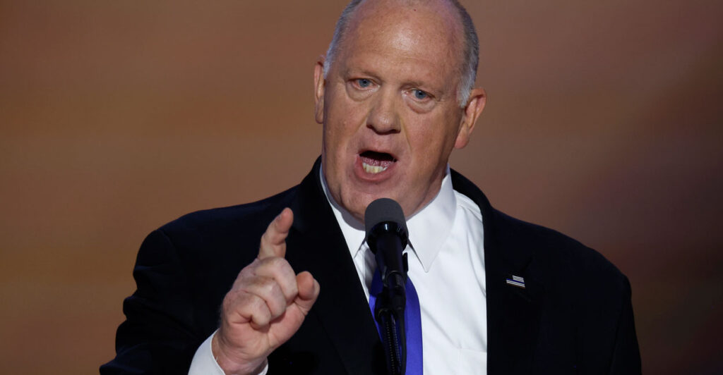 Tom Homan in a dark suit and tie speaking on stage forcefully with his finger pointing forward