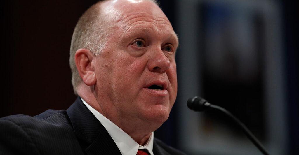 Tom Homan speaks into a microphone while seated and wearing a dark suit and red tie.