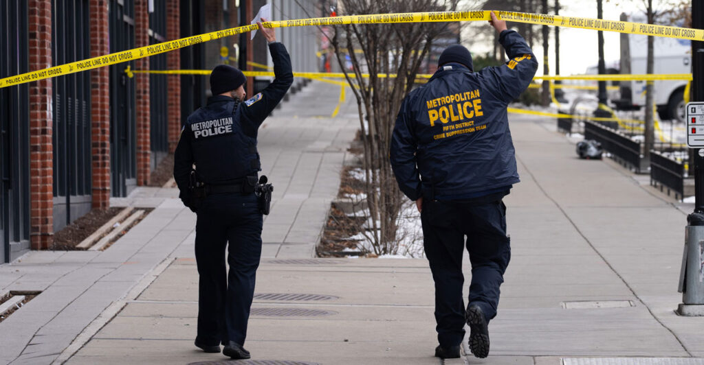 two police officers in Metropolitan Police Department jackets walk down a sidewalk with their backs to the camera holding up police caution tape as they walk under it to a crime scene