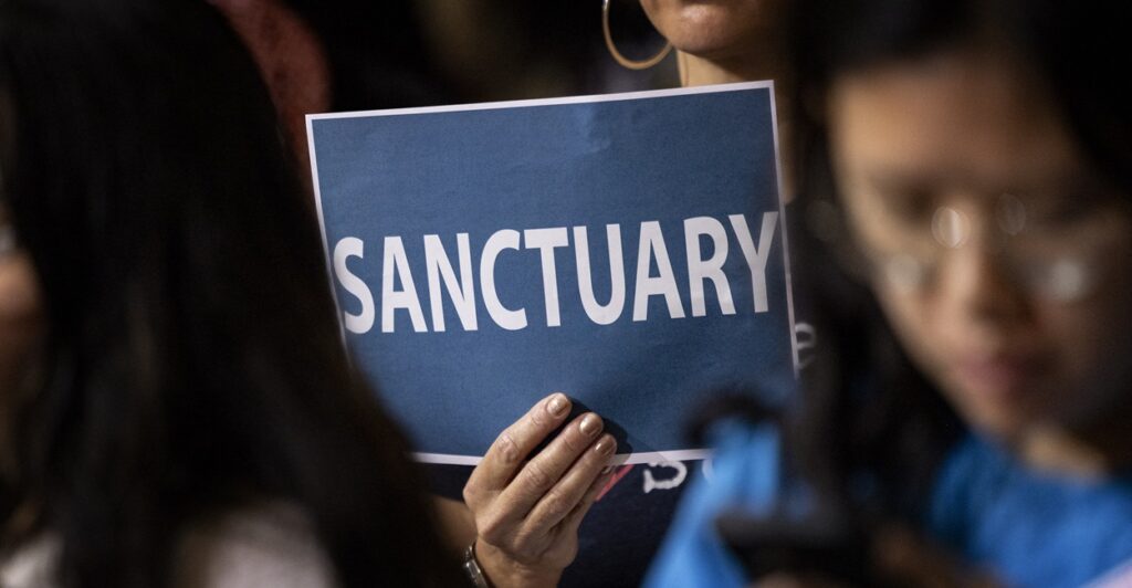 A woman holds up a sign that says "SANCTUARY" at a meeting.