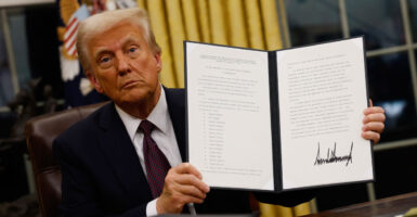Donald Trump in a black suit and red tie sitting at his desk in the oval office holding up an executive order he just signed