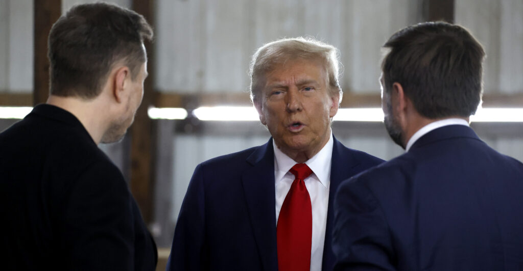 Donald Trump in a suit speaks with J.D. Vance and Elon Musk