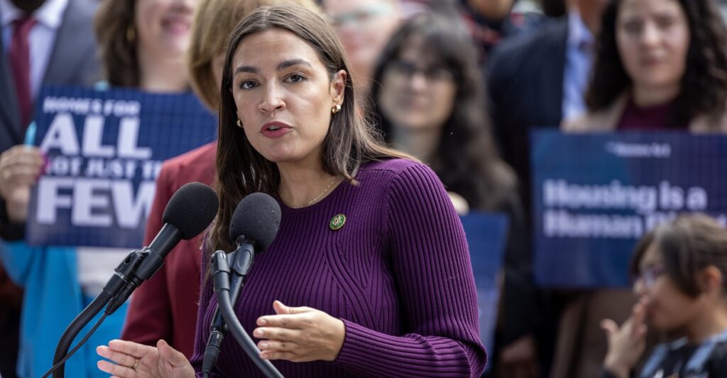 Alexandria Ocasio-Cortez stands in fron of a microphone surrounded by reporters answering questions.