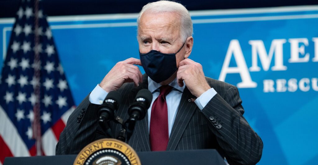 Joe Biden stands and adjusts his mask while talking during a press conference.