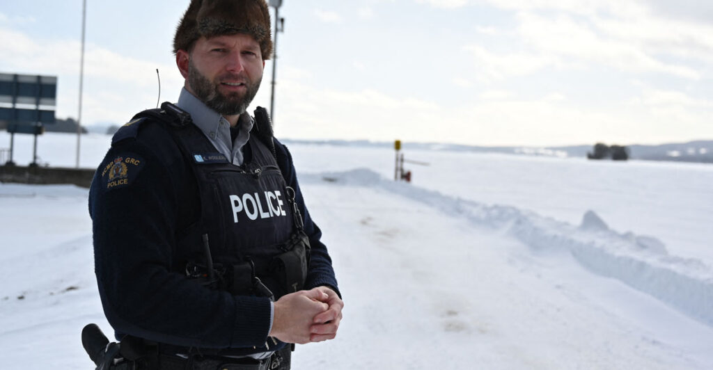 A Royal Canadian mounted police officer standing next to a frozen, snow-covered lake near the U.S. border