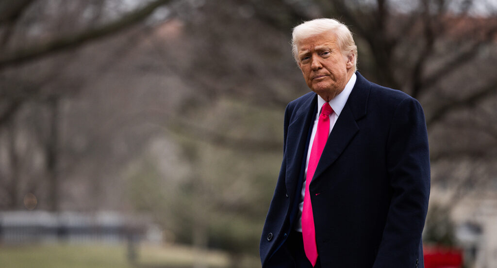 Donald Trump in a blue suit with a red tie walks outside