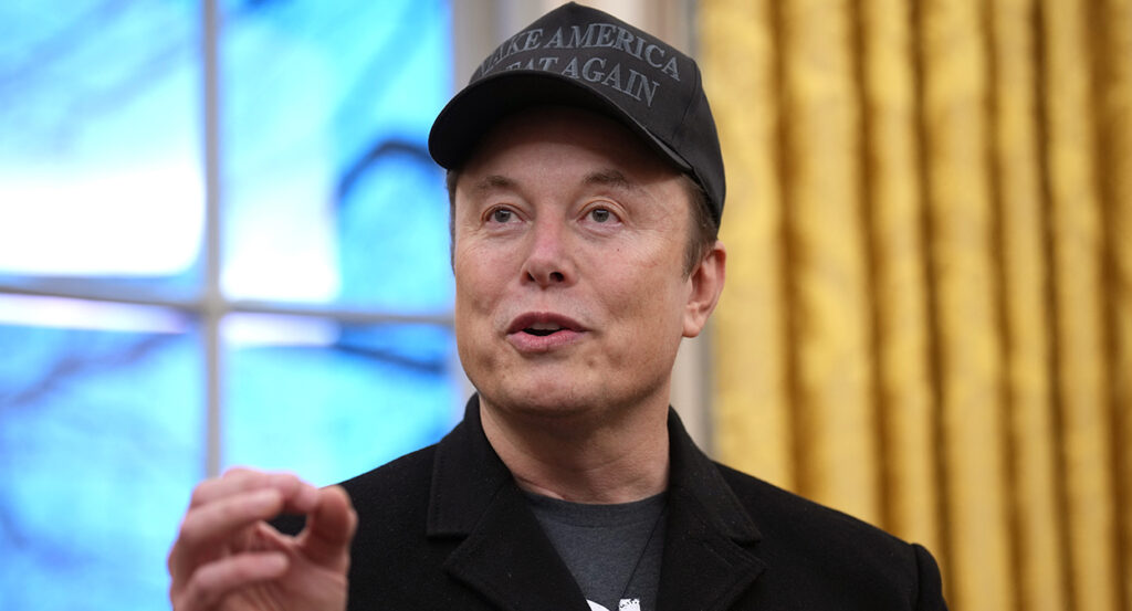 Elon Musk gestures while wearing a Make America Great Again hat in a black sportcoat and a t-shirt.