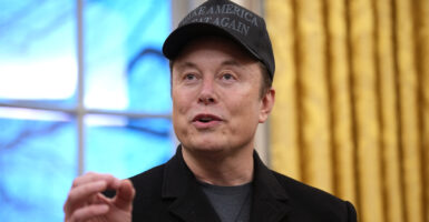 Elon Musk gestures while wearing a Make America Great Again hat in a black sportcoat and a t-shirt.