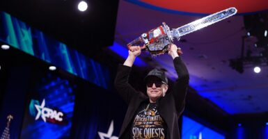 Elon Musk holds a chain saw above his head while wearing a baseball cap and sun glasses.