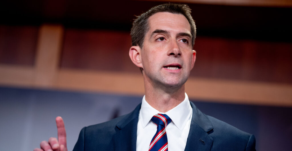 Tom Cotton stands pointing, wearing a dark grey suit and stiped necktie.
