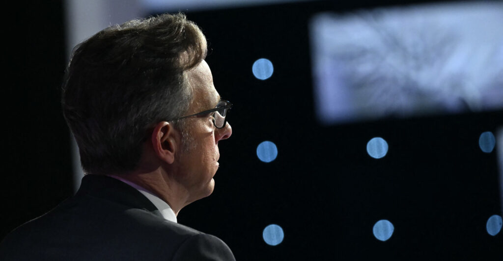 Jake Tapper sits turned away in a darkened room wearing a slate gray suit and his glasses.