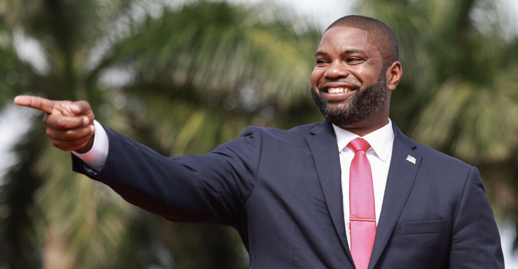 Byron Donalds wears a dark grey suit and red necktie as he smiles.