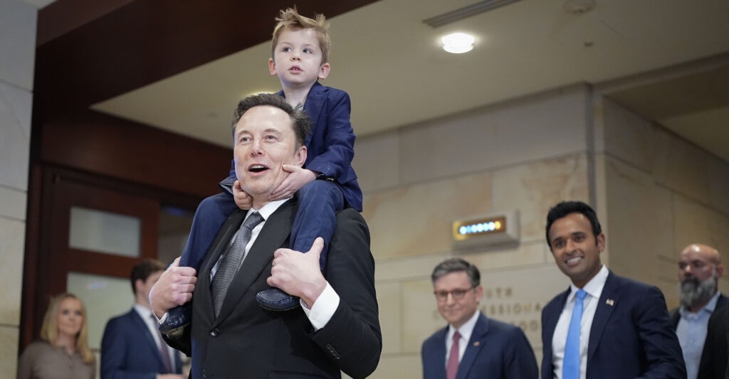 Elon Musk, dressed in a black suit, carries his young son in a blue suit on his shoulders.