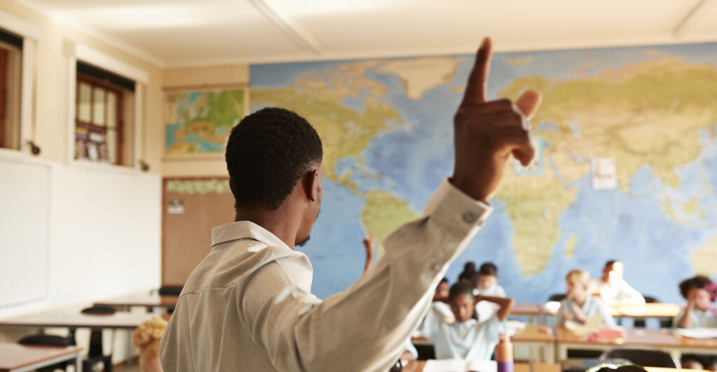 A black male teacher stands in front of a classroom wearing a white dress shirt.