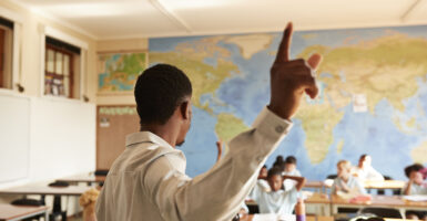 A black male teacher stands in front of a classroom wearing a white dress shirt.