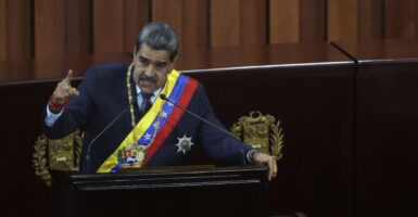 Nicolás Maduro stands in navy blue official attire including a sash across his body decorated in the colors of the Venezuelan flag.