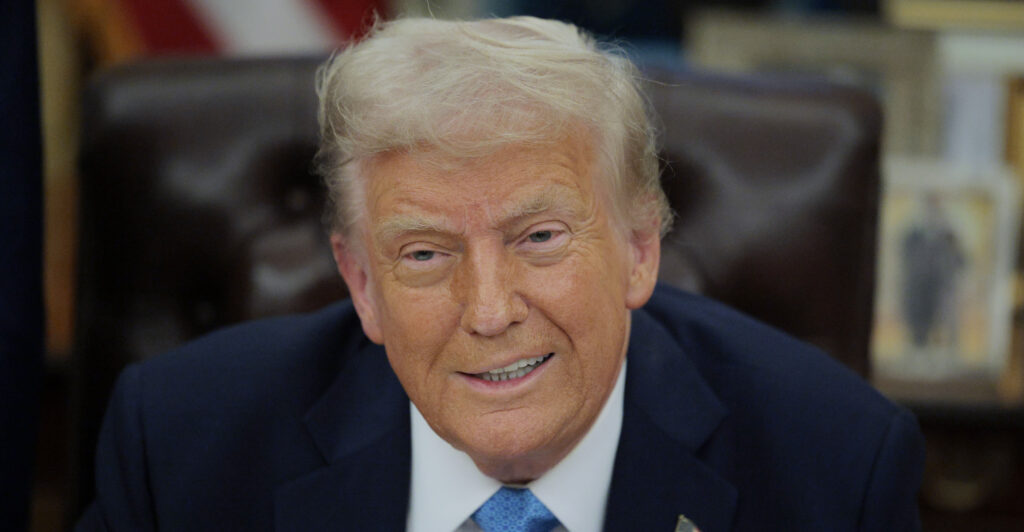 Donald Trump smiles, wearing a dark blue suit and light blue necktie.