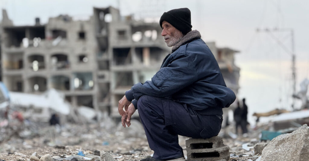 A Palestinian man sits on a gray cinderblock, wearing a black winter hat, blue winter coat, and blue pants.