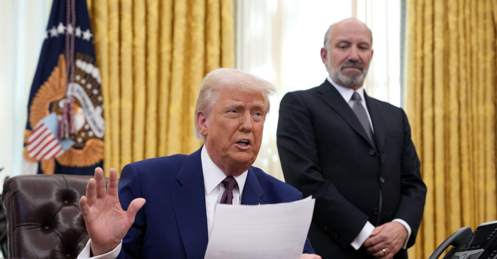 Commerce Secretary Howard Lutnick looks on as President Donald Trump sits behind the Resolute Desk in the Oval Office.