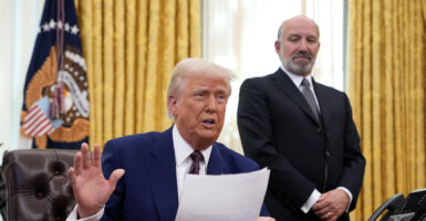 Commerce Secretary Howard Lutnick looks on as President Donald Trump sits behind the Resolute Desk in the Oval Office.