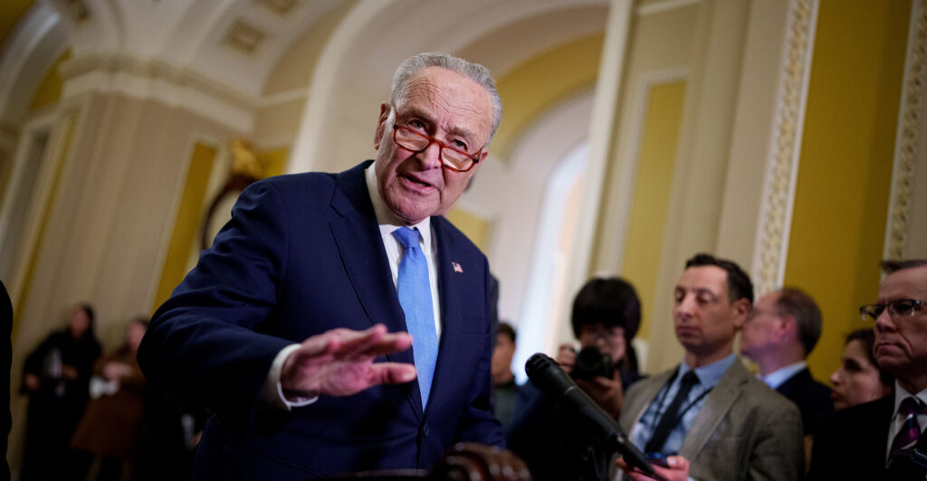 Chuck Schumer stands wearing a dark blue suit and a light blue necktie.