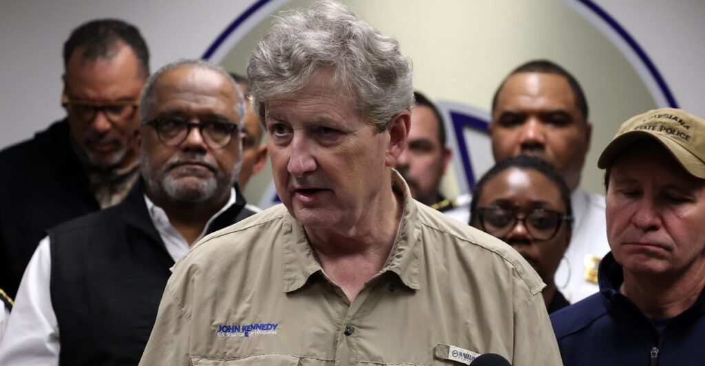 John Kennedy stands answering reporters' questions as administrators stand behind him.