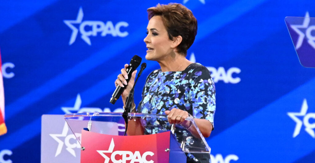 Kari Lake speaking at a podium, holding a microphone at the CPAC conference