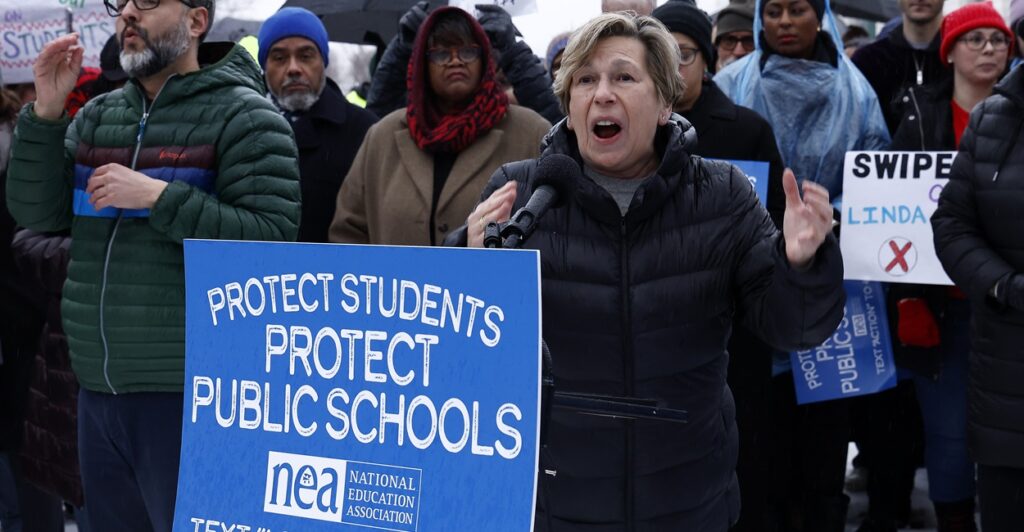Randi Weingarten is standing outside wearing a winter jacket as she shouts during a protest near a sign.