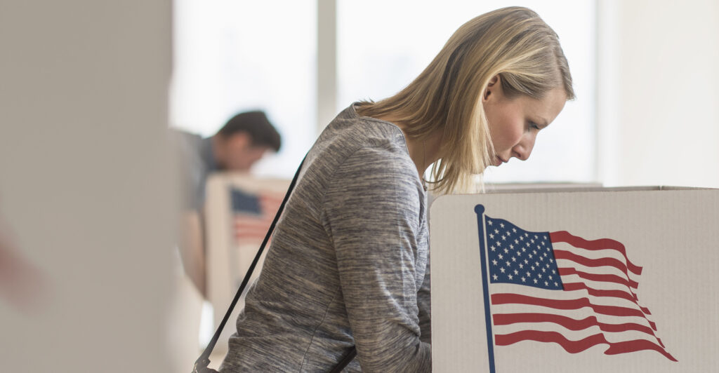 a blonde woman in a white outfit voting at a voting station with an American flag on the side of it