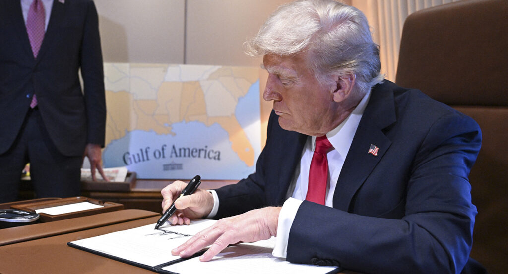 Donald Trump in a blue suit with a red tie signs an executive order with a map reading "Gulf of America" in the background.