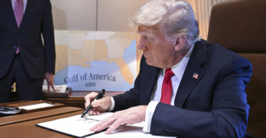 Donald Trump in a blue suit with a red tie signs an executive order with a map reading "Gulf of America" in the background.