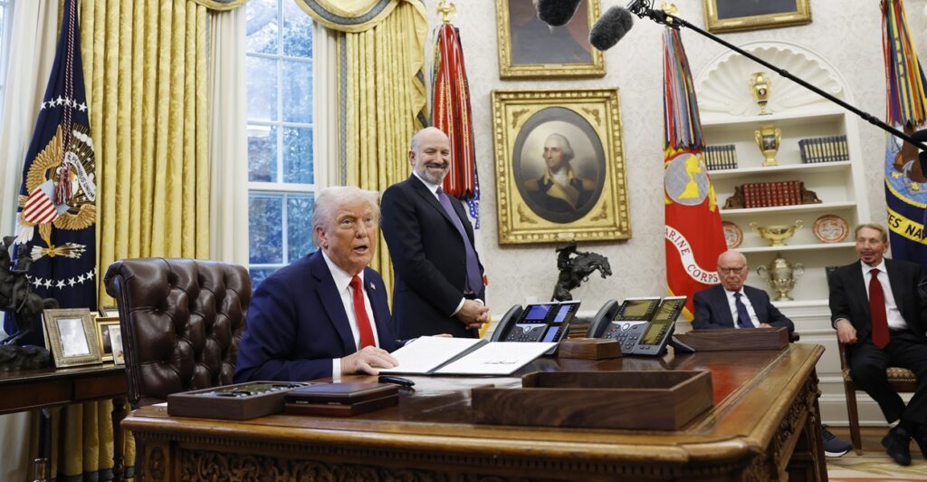 With microphones around him, Donald Trump sits at his desk and talks to reporters.