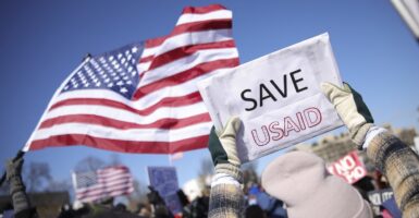 A pair of hands hold up a sign that states "SAVE USAID" as an American flag waves in the background.
