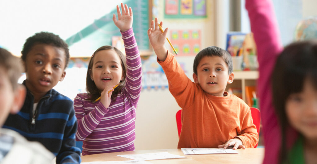 Elementary school students raise their hands as if to offer to answer a teacher's question.
