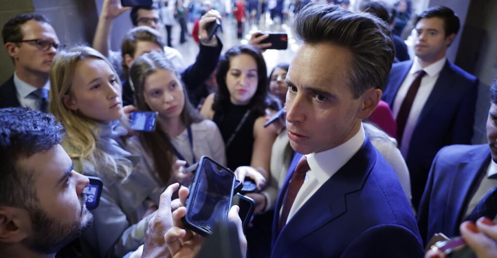 Josh Hawley is standing outdoors surrounded by reporters putting their microphones in front of his face.