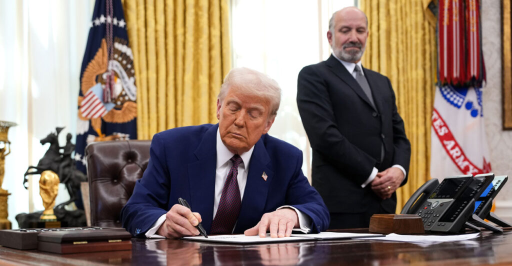 Donald Trump, in a dark suit and a tie, sits at his oval office desk looking down and signing an executive order while Howard Lutnick, in a dark suit and tie, stands behind him looking on.