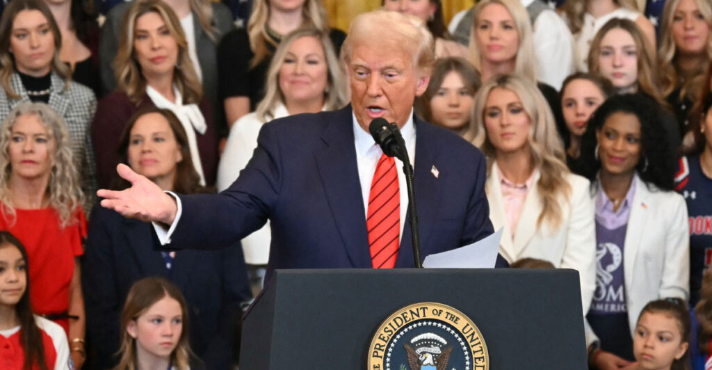 Donald Trump in a blue suit and red tie at the podium gesturing toward the crowd with several women standing behind him