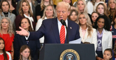 Donald Trump in a blue suit and red tie at the podium gesturing toward the crowd with several women standing behind him
