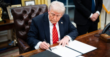 Donald Trump in a blue suit and red tie at the oval office desk signing an executive order